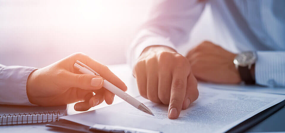 Hände und Oberkörper von zwei Männern in Businesshemden, die mit Finger und Stift auf Papiere zeigen, die auf dem Tisch liegen