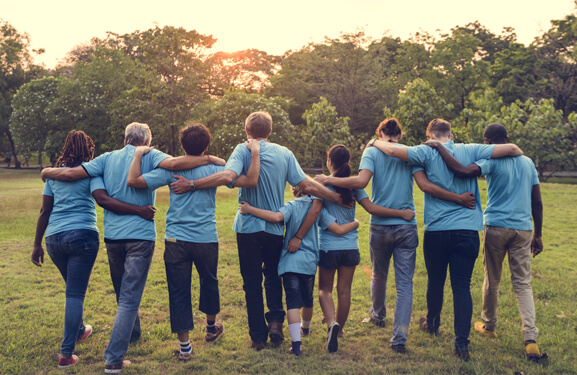 Eine bunt gemischte Gruppe von Personen mit blauen T-Shirts laufen sich gegenseitig umarmend vom Betrachter weg in Richtung eines Waldes in den Sonnenuntergang.