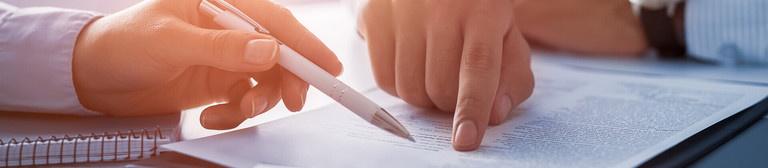 Hands and torsos of two men in business shirts pointing with finger and pen at papers lying on the table