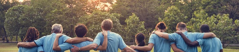 Eine bunt gemischte Gruppe von Personen mit blauen T-Shirts laufen sich gegenseitig umarmend vom Betrachter weg in Richtung eines Waldes in den Sonnenuntergang.