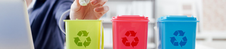 Three mini-waste bins in green, red and blue and featuring the recycling symbol standing on an office desk. Behind them, a smiling woman can be seen working at a laptop, who is throwing a crumpled piece of paper into the green bin. 